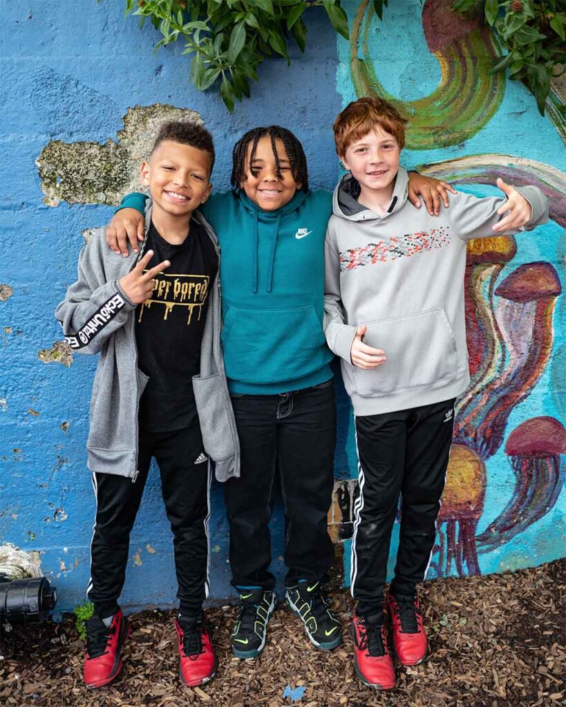 A group of three diverse fourth grade boys smile and pose. The boy in the middle has his arms over the other two boys. The boy on the left flashes a peace sign.