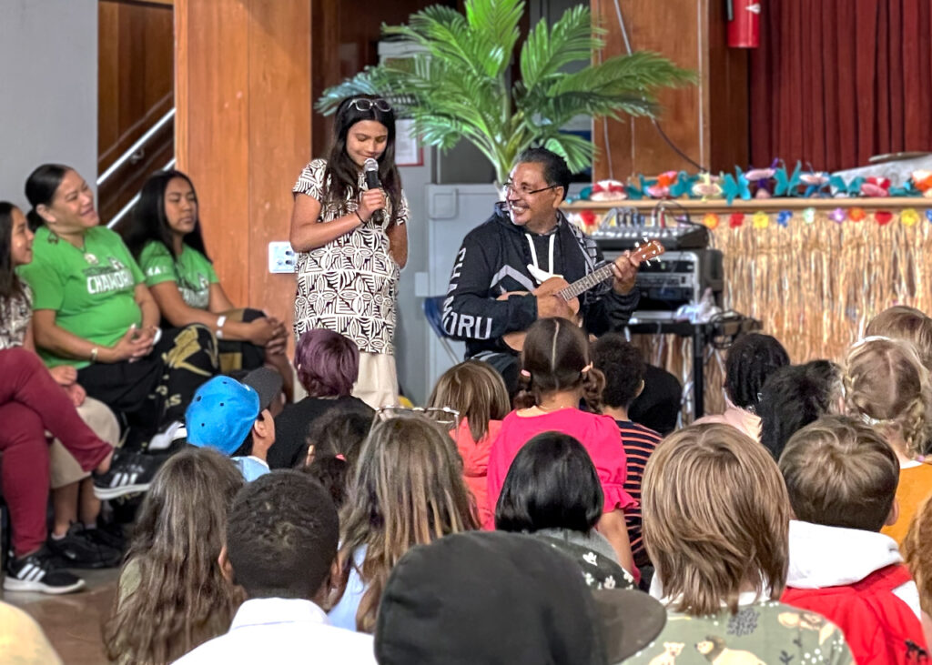 Coach Glenn plays ukulele with a student singing and kids watching during the AAPI assembly.