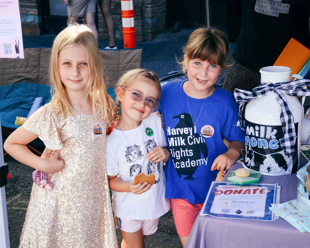 Three girls stand with a donation bucket and sign; one wears a Harvey Milk Civil Rights Academy t-shirt.