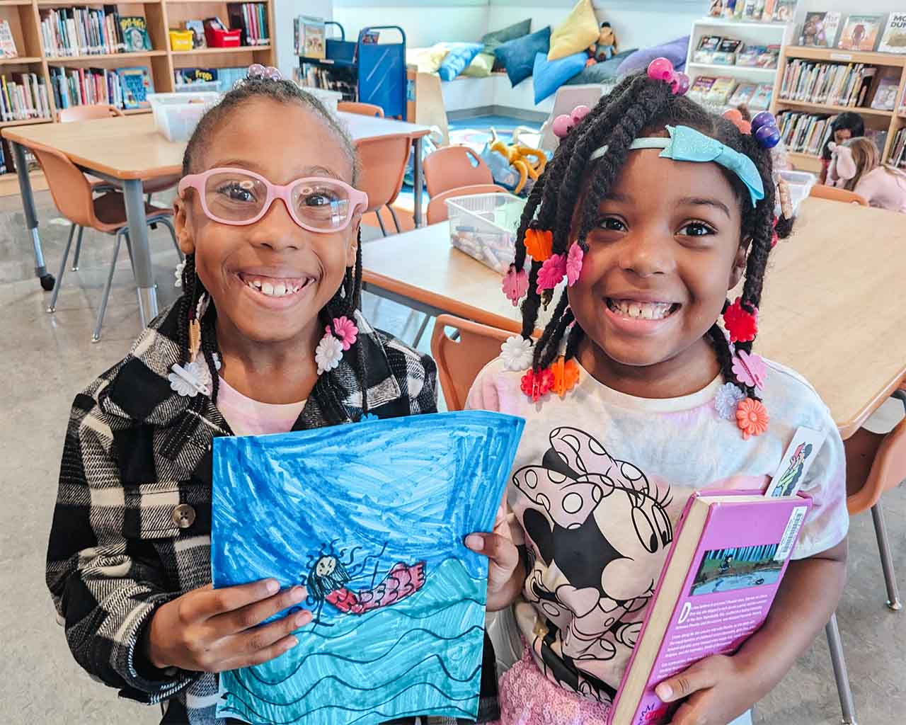 Two second grade girls hold up their drawings in the library.