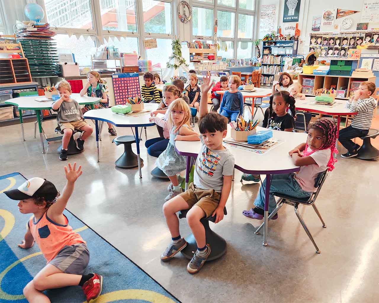 Students in a kindergarten classroom sit around circular desks and raise their hands. The classroom is filled with student artwork, posters, and school supplies.